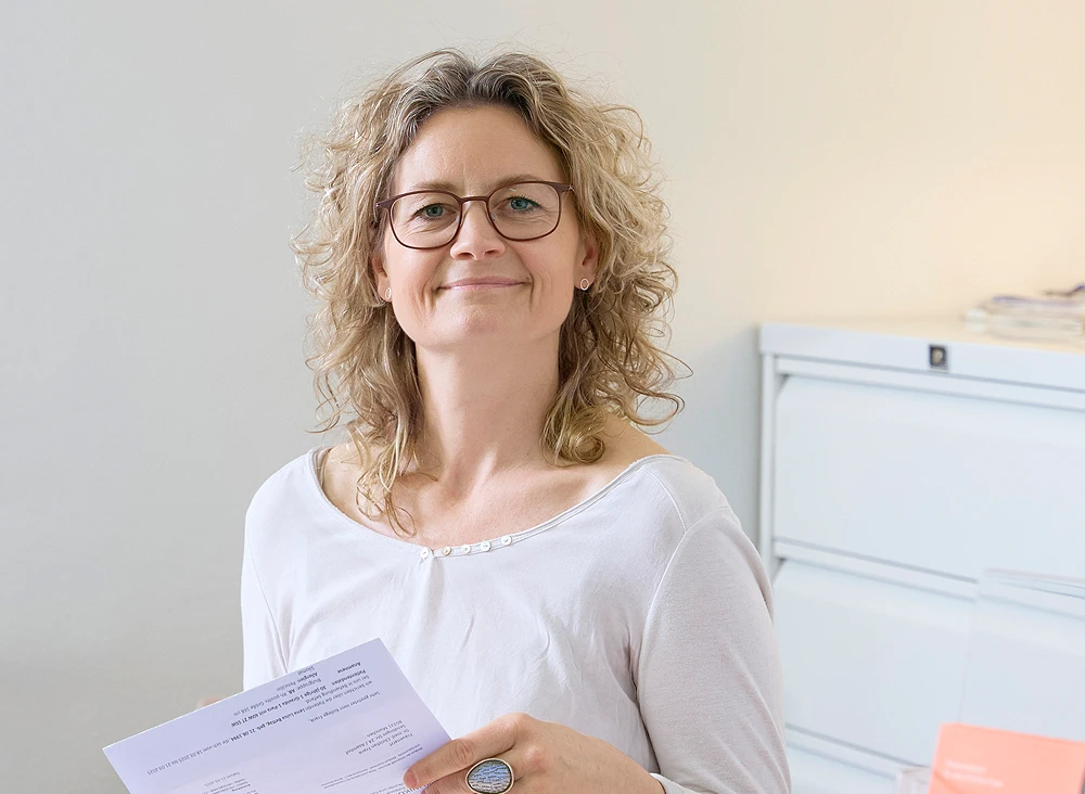 eine Frau mittleren Alters mit gelockten blonden Haaren und Brille steht vor einem Aktenschrank und lächelt freundlich in die Kamera