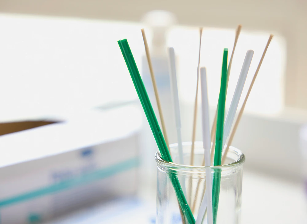 Nahaufnahme von mehreren verschiedenen Teststäbchen in einem Glas. Im Hintergrund Verpackungen von medizinischen Test-Kits