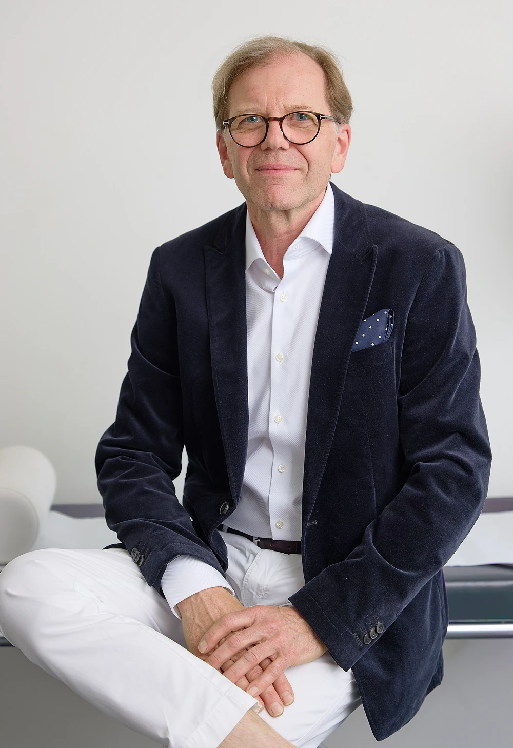 Portrait von Dr. Christian Frank, ein älterer Mann mit dünnen dunkelblonden Haaren und Brille. Er trägt eine weiße Hose, ein weißes Hemd und ein dunkles Sacko und sitzt entspannt auf der Schreibtischkante